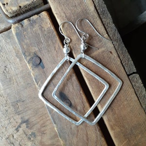 May include: Silver earrings with a hammered finish and a square shape. The earrings have a simple design with a small ball at the top of the hook.