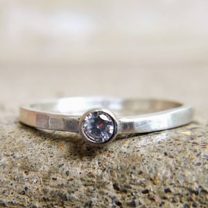 May include: A silver ring with a single, clear, round gemstone set in a bezel setting. The band is hammered and has a textured finish.