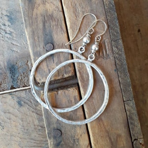 May include: A pair of silver hoop earrings with a hammered texture. The earrings are hanging from a wooden surface.