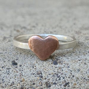 May include: A silver ring with a copper heart-shaped accent. The ring is simple and elegant.