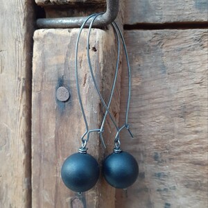 May include: A pair of black dangle earrings with round black beads. The earrings are hanging from a hook on a wooden surface.