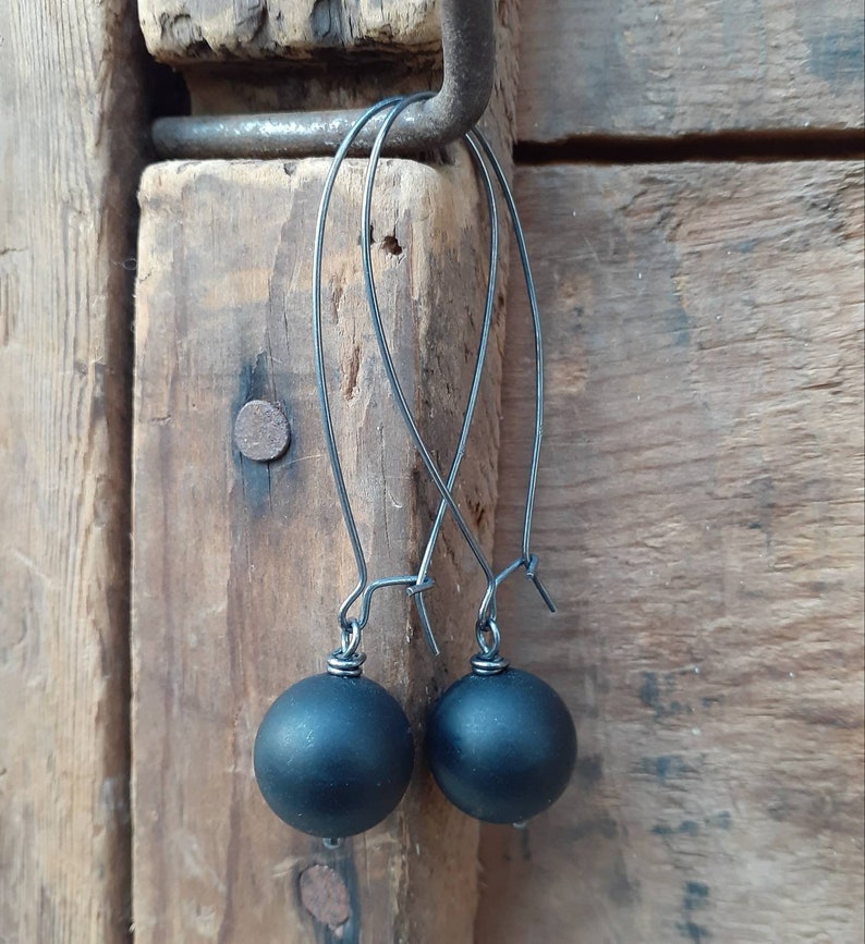 May include: A pair of black dangle earrings with round black beads. The earrings are hanging from a hook on a wooden surface.