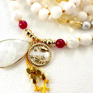 May include: A white, gold, and red beaded necklace with a gold charm featuring a zodiac symbol and a white moonstone pendant.