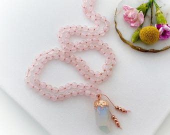 May include: A long necklace featuring light pink, translucent, spherical beads strung together. The necklace ends in a single, elongated, semi-translucent crystal pendant capped with a metallic rose gold textured top and two small beaded tassels.