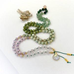 May include: A long beaded necklace featuring a gradient of green and pink gemstone beads strung on green cord. The necklace includes small gold-toned metal spacers and a teardrop-shaped pendant with a gold crescent moon charm at the end.