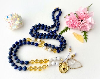 May include: A beaded necklace featuring dark blue lapis lazuli beads, translucent yellow citrine beads, and white beads. The necklace includes a gold-toned circular compass charm and a clear, triangular-shaped stone pendant with gold-toned edges.