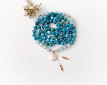 May include: A long beaded necklace featuring a variety of round blue and teal gemstone beads, interspersed with small metallic copper-colored spacers. The necklace ends in a teardrop-shaped white stone pendant with two dangling beaded strands.