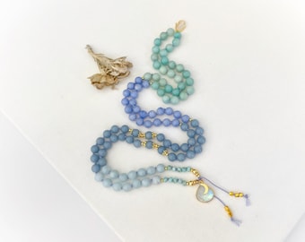 May include: A long beaded necklace featuring a gradient of round stones in shades of teal, periwinkle, and light blue. The necklace includes small gold-toned spacer beads, a teardrop-shaped pendant with a gold crescent moon, and two tassels.