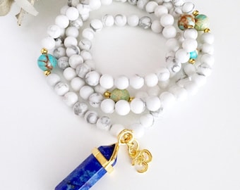 May include: A multi-strand beaded necklace featuring white and grey marbled beads, interspersed with round turquoise and brown accent beads. A gold-toned metal pendant shaped like a pointed crystal in deep blue hangs with a gold Om symbol charm.