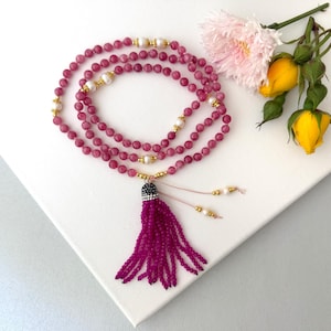 May include: A long beaded necklace featuring round magenta gemstone beads interspersed with small white pearls and gold-toned metal spacers. The necklace ends in a large, dark magenta beaded tassel with a crystal-encrusted cap and two thin beaded strands.