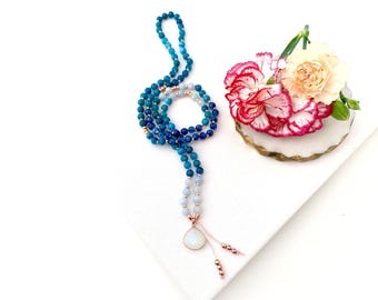 May include: A long beaded necklace featuring a gradient of blue and light blue spherical beads, accented with small metallic rose gold spacers. The necklace terminates in a teardrop-shaped pendant with a white stone and two dangling beaded strands.