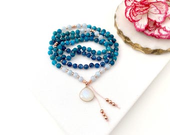 May include: A multi-strand beaded necklace featuring shades of blue and light blue gemstone beads, accented with small rose gold metallic spacers. The necklace includes a teardrop-shaped white moonstone pendant and two dangling rose gold bead ends.