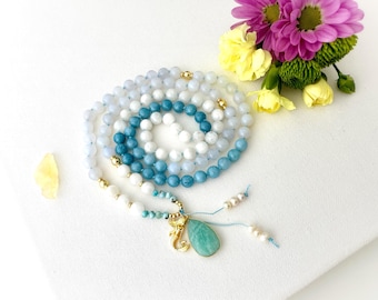 May include: A beaded necklace featuring a spiral arrangement of light blue, white, and turquoise round beads accented with small gold-toned spacers. A gold-toned mermaid charm and a teardrop-shaped green stone pendant hang from the end.
