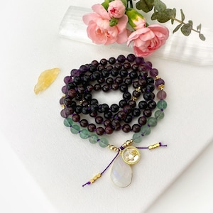 May include: A beaded mala necklace featuring dark purple, deep red, and translucent green gemstone beads strung on a purple cord. The necklace includes gold-toned metal spacers, a circular charm with a small embedded stone, and a teardrop-shaped pendant.