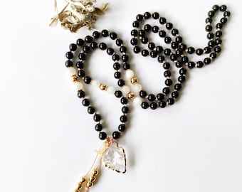 May include: A long beaded necklace featuring smooth black spherical beads, interspersed with smaller white beads and gold-toned metallic accents. A clear, jagged-edged crystal pendant hangs from the centre, accompanied by a small gold crescent moon charm.