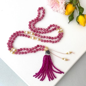 May include: A long beaded necklace featuring round magenta gemstone beads, interspersed with white pearls and small gold-toned spacers. The necklace terminates in a large, multi-strand magenta beaded tassel with a dark metallic cap.