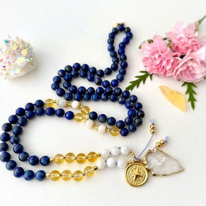 May include: A blue and yellow gemstone mala necklace with a gold compass charm and a clear quartz point. The necklace is made with lapis lazuli, citrine, and white howlite beads.