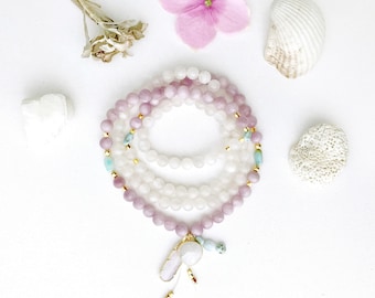 May include: A multi-strand beaded necklace featuring smooth, round light purple and translucent white beads. The necklace includes small gold-toned spacers, occasional light blue accent beads, and a central pendant with a white stone.