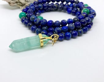 May include: A beaded necklace featuring deep blue lapis lazuli spheres interspersed with occasional green marbled beads. A translucent, pale green, hexagonal crystal point pendant is attached to the strand with a gold-toned metal bail.