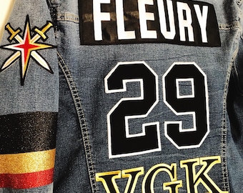 Puede incluir: Una chaqueta vaquera azul con un parche "FLEURY 29" en blanco y negro y un parche "VGK" dorado en la espalda. La manga izquierda tiene un parche a rayas rojo, dorado y negro con una estrella y espadas cruzadas.