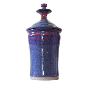 May include: A blue ceramic jar with a lid. The jar has a speckled finish and a flared shape. The lid is a darker blue with a red accent. The jar is a decorative item.