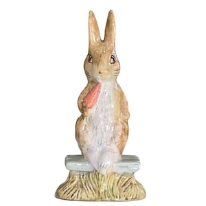 May include: A ceramic figurine of a brown rabbit holding a carrot. The rabbit is sitting on a light blue bench with a straw base. The rabbit has long ears and a white chest. The carrot is orange.
