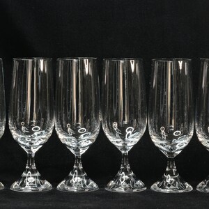 May include: Six clear, stemmed champagne flutes with vertical etched lines. The glasses have a tulip shape and sit on a decorative base. The background is solid black.