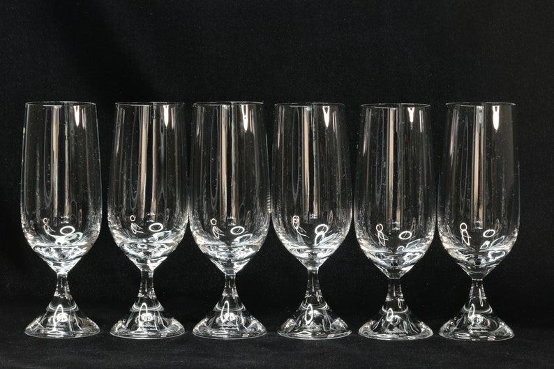 May include: Six clear, stemmed champagne flutes with vertical etched lines. The glasses have a tulip shape and sit on a decorative base. The background is solid black.
