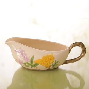 May include: Cream-colored ceramic gravy boat with a floral design. The boat features a yellow flower and purple flowers with green leaves. The handle has a twisted, brown design.