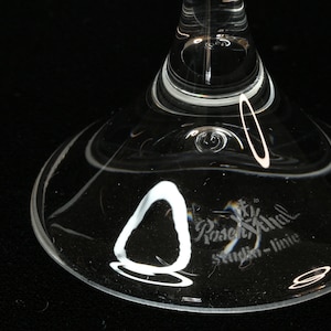 May include: Close-up of the base of a clear glass stemware. The glass has a wide, rounded base with a delicate, curved design. The text "Rosenthal studio-linie" is etched on the glass.