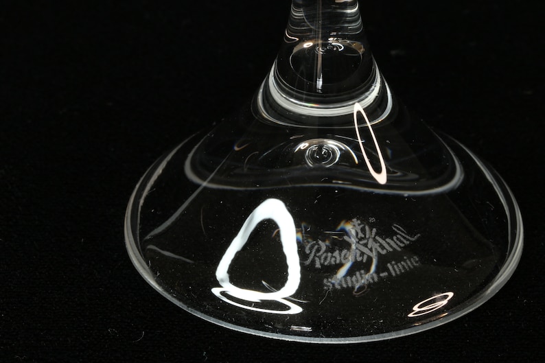 May include: Close-up of the base of a clear glass stemware. The glass has a wide, rounded base with a delicate, curved design. The text "Rosenthal studio-linie" is etched on the glass.