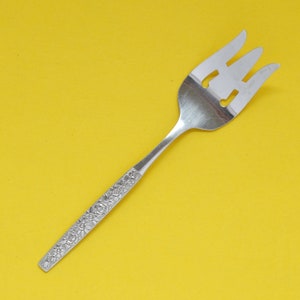May include: A silver serving fork with three tines and a floral pattern on the handle. The fork is made of stainless steel and is designed for serving food. The tines have cut-out designs. The background is a solid yellow.