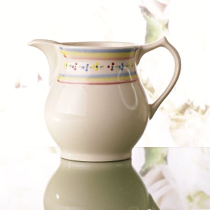 May include: Cream-colored ceramic pitcher with a curved handle and spout. The pitcher features a decorative band of horizontal stripes in yellow, blue, and red, accented with small, colorful dots. The pitcher is reflected on a glossy surface.
