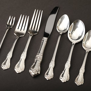 May include: A set of ornate silverware, including forks, a knife, and spoons, arranged on a dark surface. The silverware has a shiny, silver finish with detailed floral patterns on the handles. The set includes three forks, a knife, and three spoons.