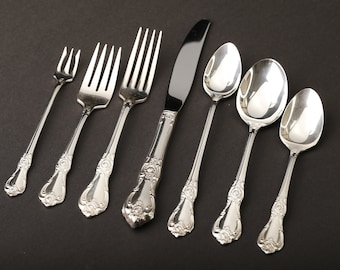 May include: A set of ornate silverware, including forks, a knife, and spoons, arranged on a dark surface. The silverware has a shiny, silver finish with detailed floral patterns on the handles. The set includes three forks, a knife, and three spoons.