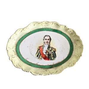 May include: Oval decorative tray with a portrait of a man in military attire. The tray has a scalloped edge with a cream-colored border and a green inner oval. The portrait features a man with a red sash and gold accents.