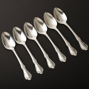 May include: Six silver spoons arranged diagonally on a black surface. Each spoon has a rounded bowl and a decorative handle with floral detailing. The spoons are reflective, showing the surrounding environment.
