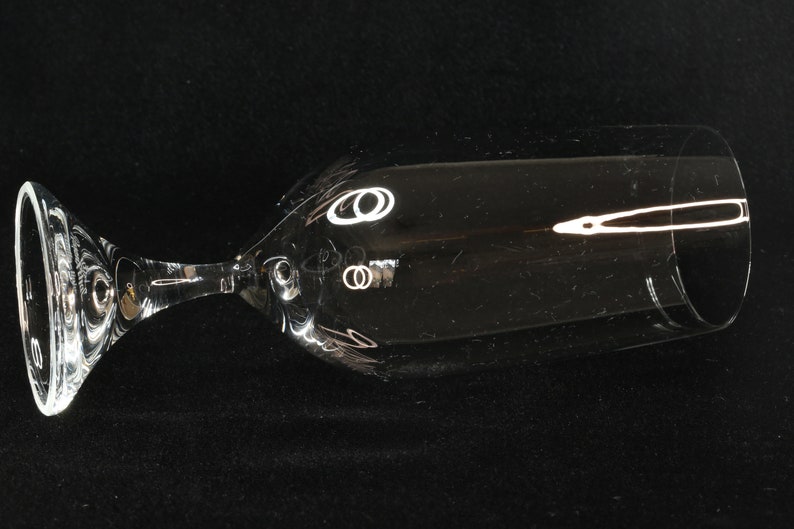 May include: A clear, stemmed champagne glass with a wide bowl and a slender stem. The glass has a decorative design near the top of the bowl. The glass is lying on its side against a black background.