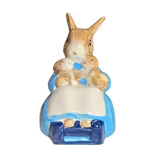 May include: A ceramic figurine of a rabbit wearing a blue dress and holding three smaller rabbits. The figurine has a white apron and a blue seat. The rabbit has brown and tan fur.