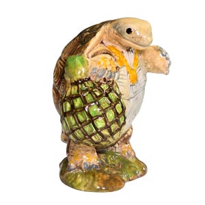 May include: A ceramic turtle figurine with a detailed shell, holding a green bag with a diamond pattern. The turtle has a yellow necklace and is standing on a green base. The figurine is painted in earth tones.
