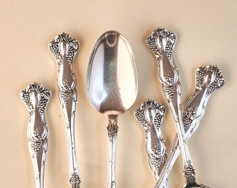 May include: A collection of ornate silver spoons arranged on a light beige surface. The spoons feature detailed grape cluster designs on the handles and decorative elements at the base of the spoon bowls. The spoons vary in size and shape, reflecting a formal dining setting.