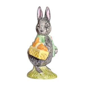 May include: A ceramic figurine of a gray rabbit wearing a green jacket and holding a yellow basket filled with orange carrots. The rabbit stands on a patch of green grass, with pink details on its ears.