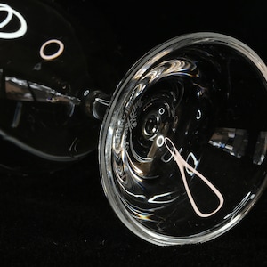 May include: A clear, stemmed wine glass with a wide bowl, resting on its side. The glass reflects light, creating bright highlights and a soft pink design on the bowl. The background is black.