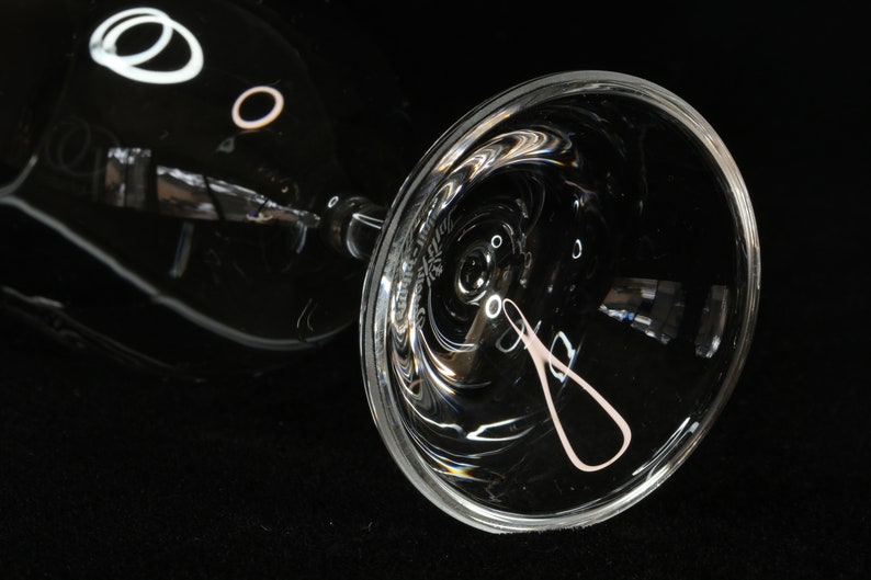 May include: A clear, stemmed wine glass with a wide bowl, resting on its side. The glass reflects light, creating bright highlights and a soft pink design on the bowl. The background is black.