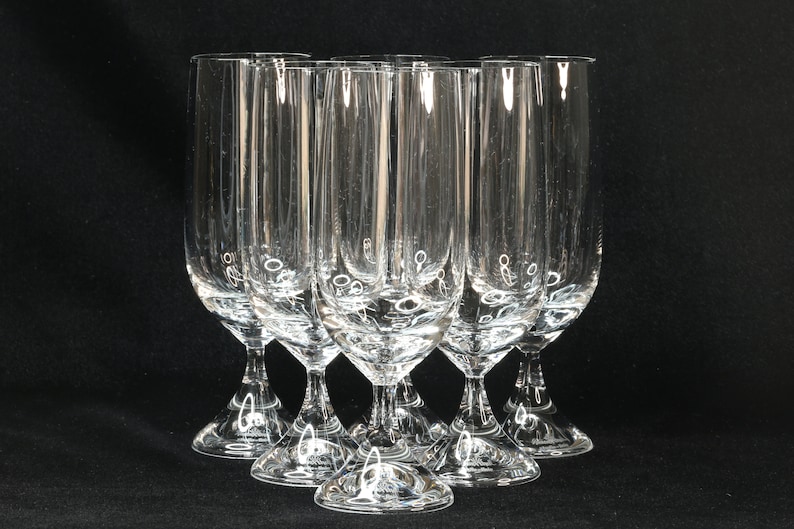 May include: Six clear, stemmed champagne flutes arranged in a semi-circle. The glasses have a tall, slender bowl with vertical lines and a flared base. The background is a solid black.
