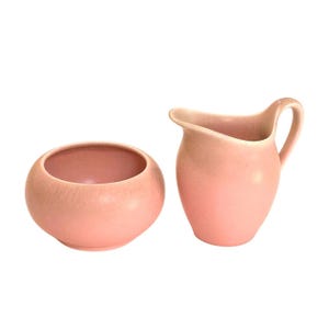 May include: A pink ceramic creamer pitcher with a handle and a matching round sugar bowl. The set is a soft, pastel pink color and has a smooth, glossy finish. The sugar bowl has a wide opening.