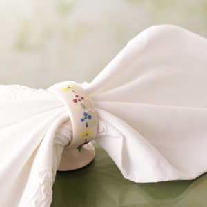 May include: A cream-colored ceramic napkin ring with a floral design of yellow, red, and blue flowers. The ring holds a white cloth napkin. The napkin ring is on a green surface.