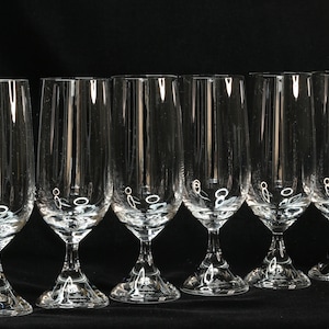 May include: Six clear champagne flutes with a tulip-shaped bowl and a long stem. The glasses are made of clear glass and are arranged in a row against a black background. The stem has a decorative design.