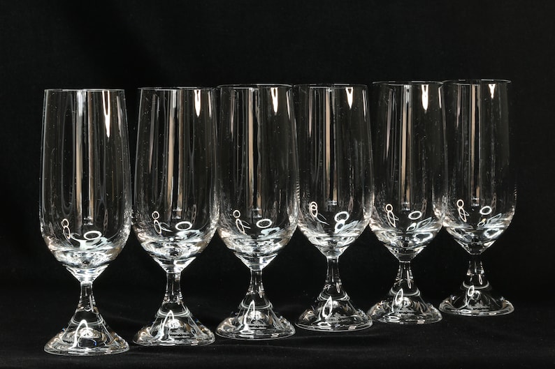 May include: Six clear champagne flutes with a tulip-shaped bowl and a long stem. The glasses are made of clear glass and are arranged in a row against a black background. The stem has a decorative design.