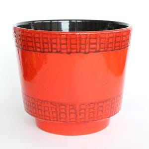 Puede incluir: Una maceta de cerámica de color rojo brillante con acabado lustroso e interior oscuro. El exterior presenta dos bandas horizontales con un patrón texturizado de rejilla en un tono rojo más oscuro que rodea el cuerpo del recipiente cilíndrico.